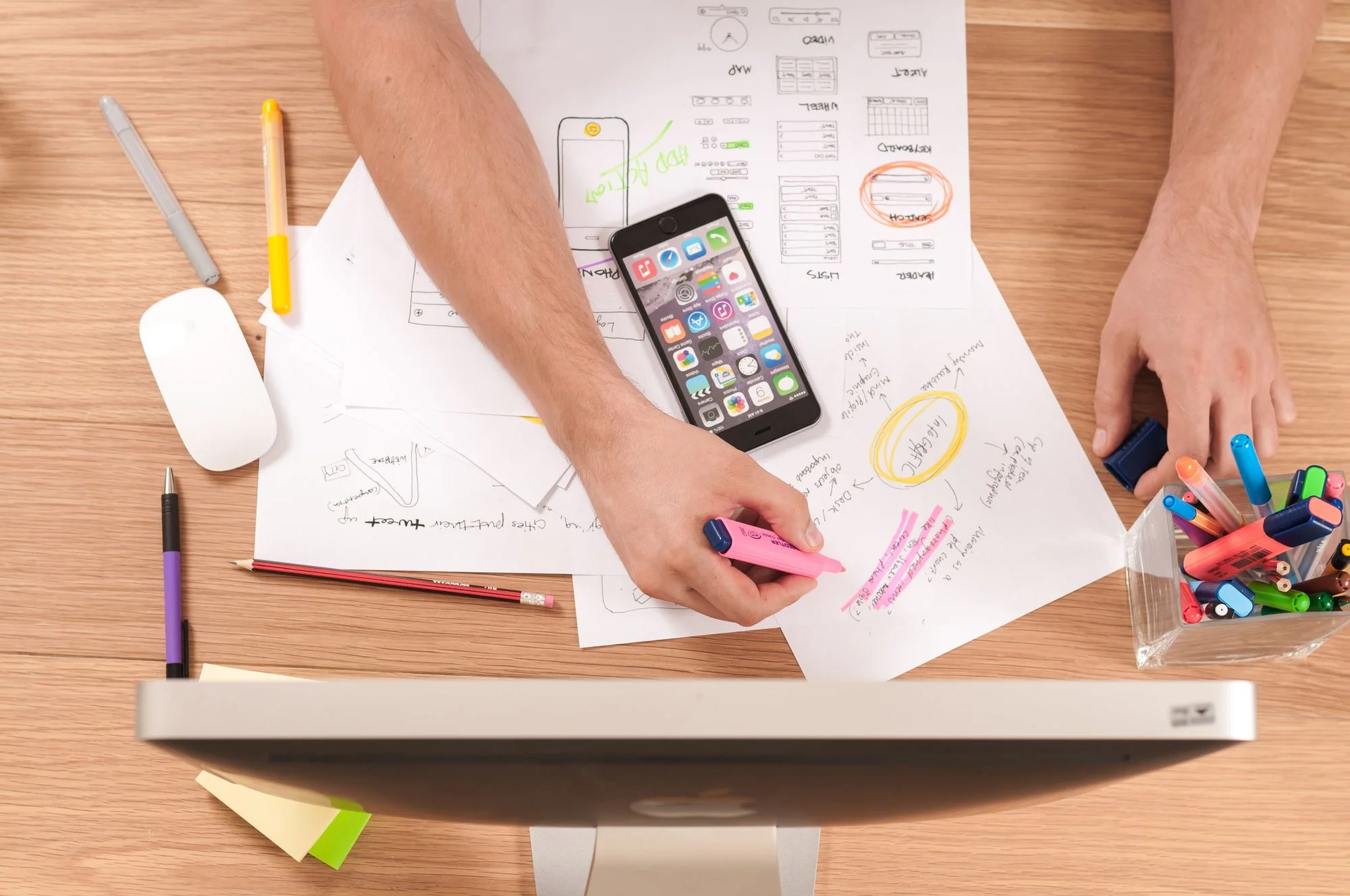 a view from above a desk with a monitor, a mouse, scattered papers, a mobile phone and a pen box on it and a person working