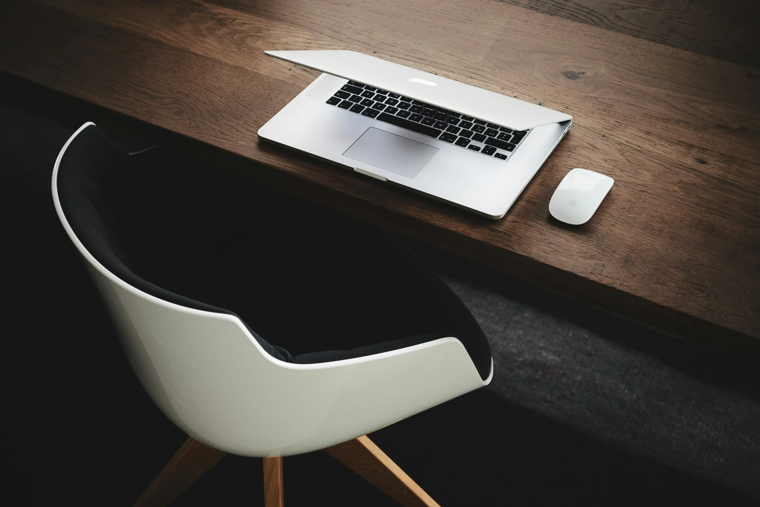 A laptop on a dark desk with a white wireless mouse and a modern white chair in front.